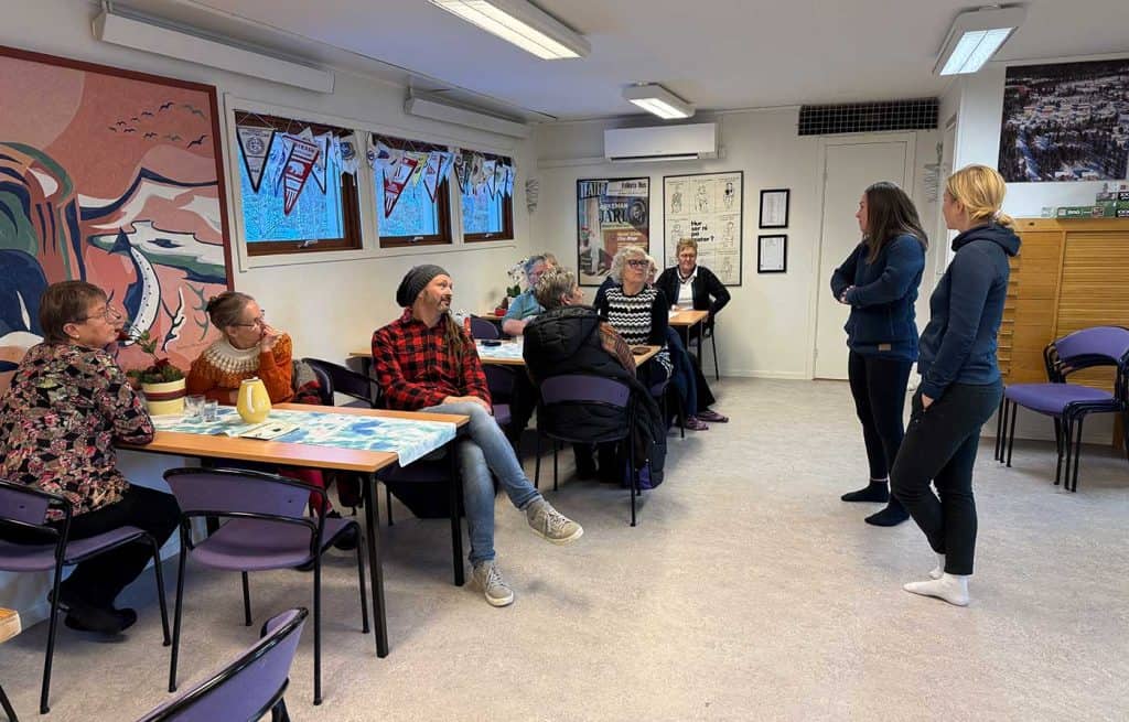 Översiktsbild i en lokal med bord längs ena väggen. Det sitter folk vid alla borden och deras uppmärksamhet är riktad mot två kvinnor som står upp framför borden.