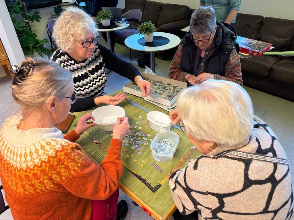 Fyra kvinnor sitter runt ett litet bord och lägger pussel. Bilden är taget snett uppifrån, och bordet är täckt av pusselbitar.