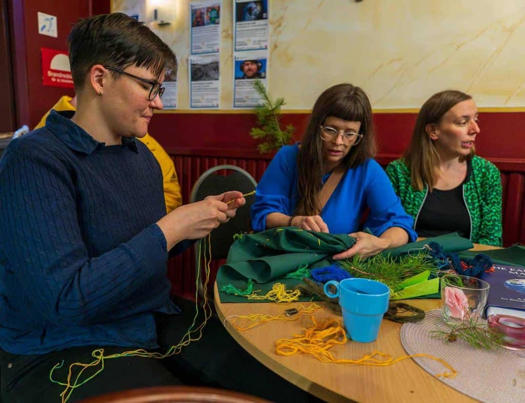 Tre kvinnor sitter vid ett bord och jobbar med ett textilt konstverk som ska symbolisera den biologiska mångfalden.