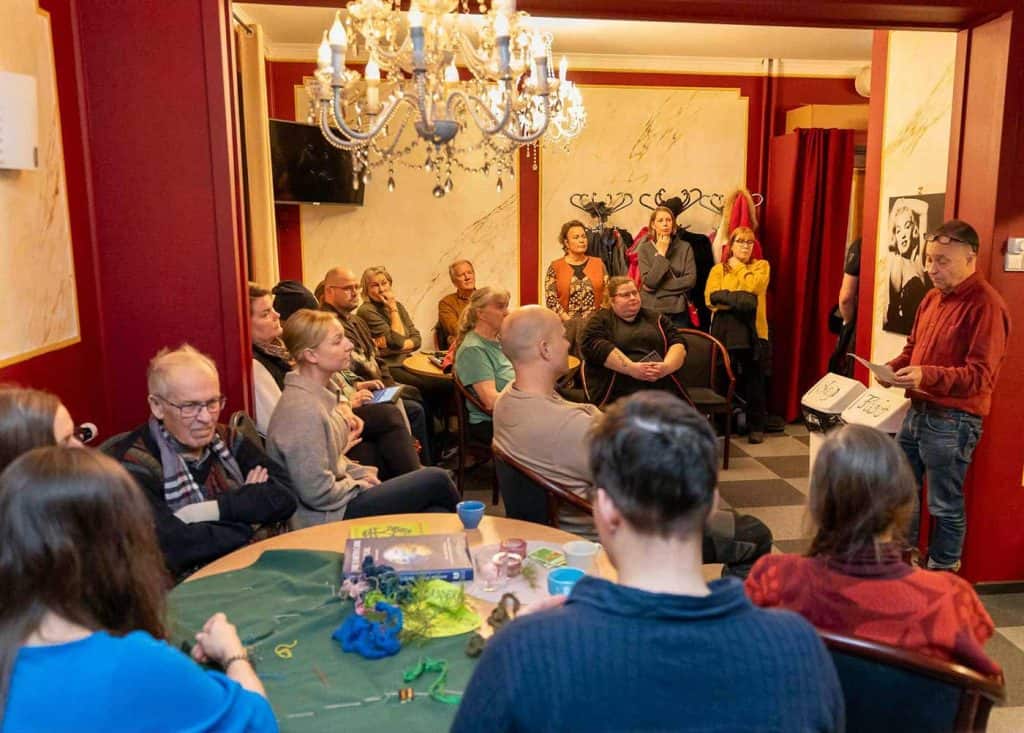 Översiktsbild i en lokal som inte är så stor (bions foaje). Ett tjugotal människor syns i bild, de sitter och står, och har uppmärksamheten riktad mot en man som läser ur ett papper han har i handen.