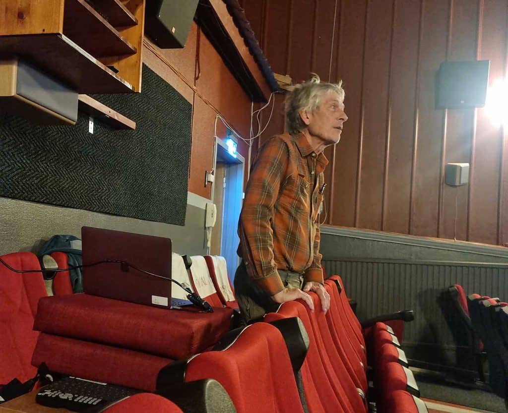 En man står längst bak i en biosalong. Han håller händerna på ryggstödet framför sig och bakom sig har han en dator med sladd. 