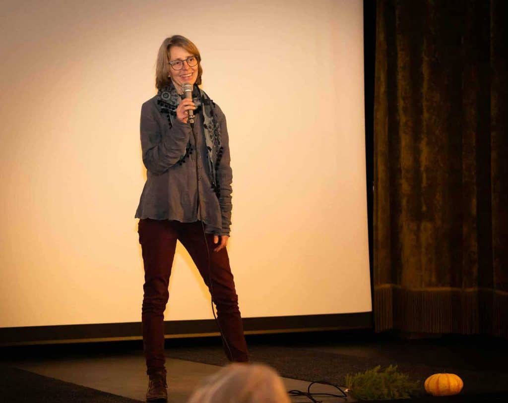 En kvinna står på en scen framför en bioduk. I sin hand håller hon en mikrofon.