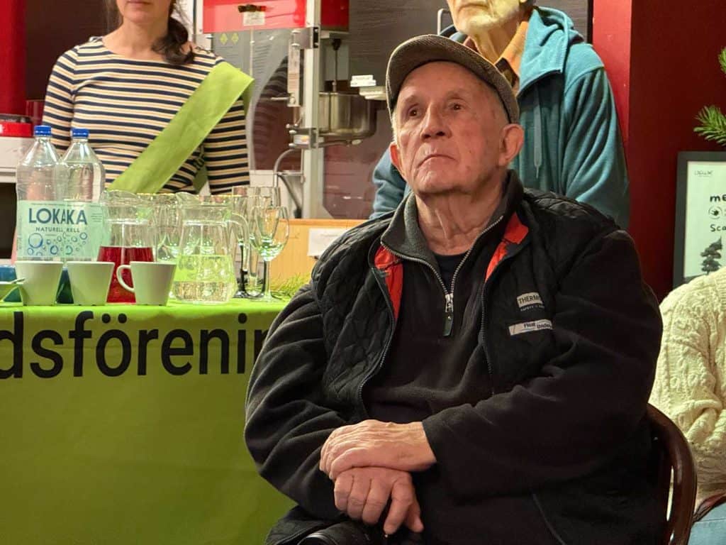 Bilden visar överkroppen på en man, sittande på en stol. Bakom mannen en bänk med glas och vattenflaskor. Bänken är täckt med en duk, med texten Naturskyddsföreningen.