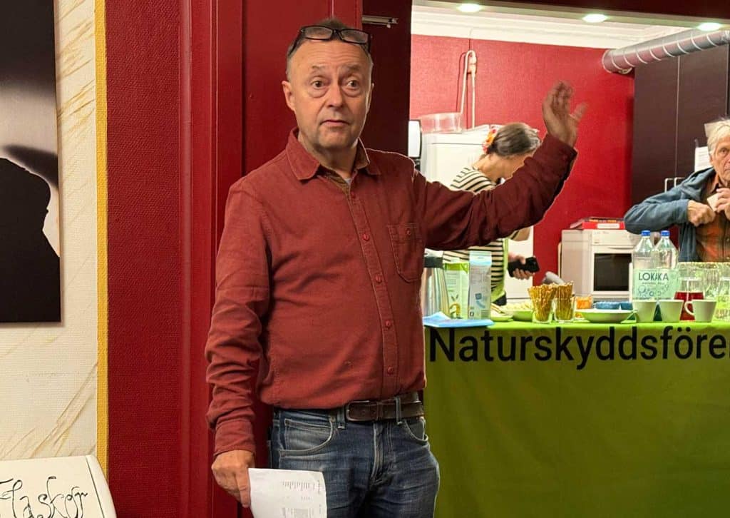 En man står och pratar och gestikulerar. I ena handen håller han ett papper. Bakom honom skymtar delar av en kvinna och en bänk med glas och vattenflaskor.