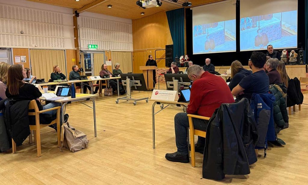 Bild på en möteslokal. Mötesdeltagarna sitter vid bord, vända mot varandra i en fyrkant med en öppen yta i mitten. I bakgrunden finns två talarstolar, och det står en person i vardera talarstolen.