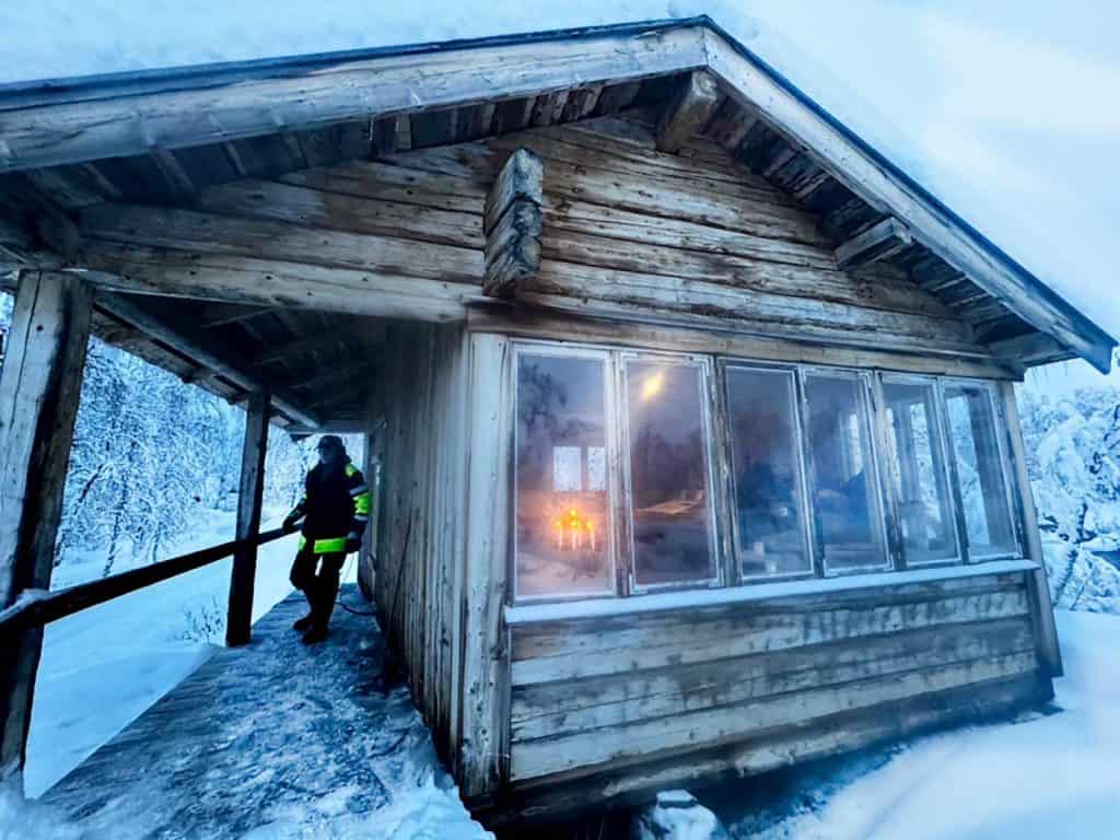 Ett timmerhus i snöigt landskap. På bron står en man i täckjacka. Genom fönstret ser man ljus brinna inne i stugan, och anar konturerna av människor.