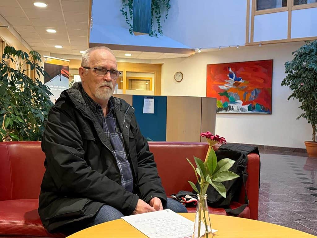 Man med skägg, glasögon och ytterkläder på sitter på kanten av en soffa i en stor entré. Bakom honom skymtar glasdörrar och bakom den en korridor med kontorsdörrar. På bordet framför mannen ligger ett papper med handskrivna anteckningar, och det finns en vas med en tygblomma i. 