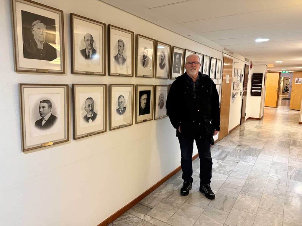 En man med skägg och glasögon, ståendes i en korridor. Bakom honom syns öppna och stängda kontorsdörrar. På väggen intill honom hänger tavlor med porträttfoton.