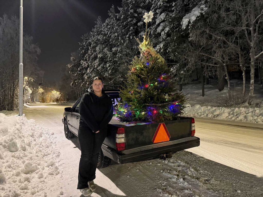 En svart EPA fotad bakifrån. På flaken står en julgran med färgglad belysning. Bredvid EPA:n står en tjej i svarta kläder. Hon ser glad ut. På EPA:ns bakskärm sitter en röd triangelformad skylt som betyder "Långsamtgående fordon".