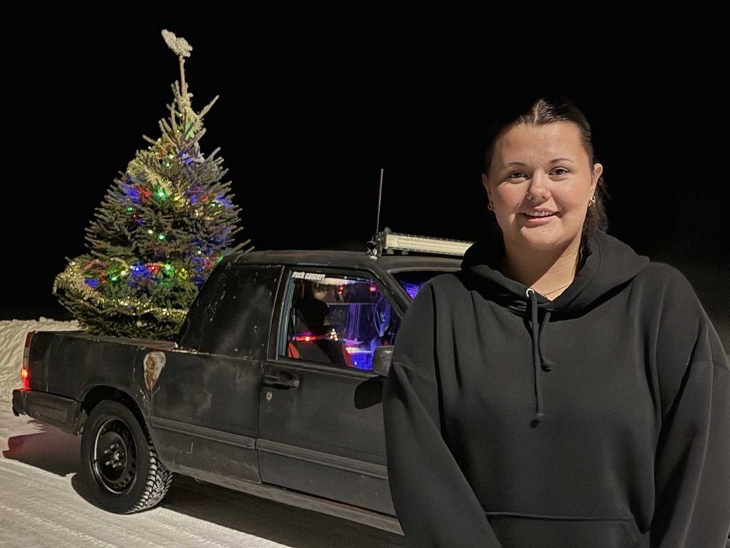 Porträttbild av en leende svartklädd tjej. Bakom henne i mörkret står en svart EPA med en ljusprydd julgran på flaket.